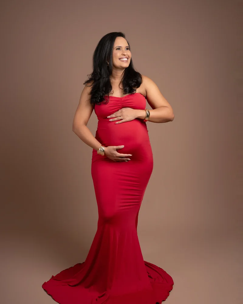 Gestante em vestido vermelho longo com as mãos na barriga, ensaio em estúdio